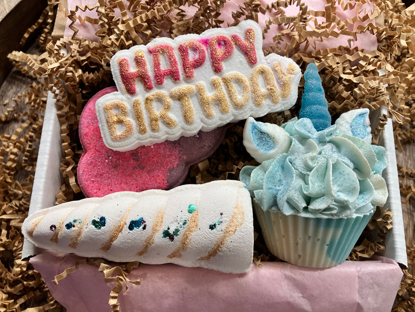 The products described previously are all arranged inside a box and cushioned with brown shred. They are arranged in the following order: The "Happy Birthday" bath bomb is on top of the playful pink and purple cloud and above the magical unicorn. The soap unicorn is on the far right of the box, beside the playful pink cloud and elegant unicorn horn bath bombs. The elegant unicorn horn is in the far right hand corner of the box with the playful pink and purple cloud above it.