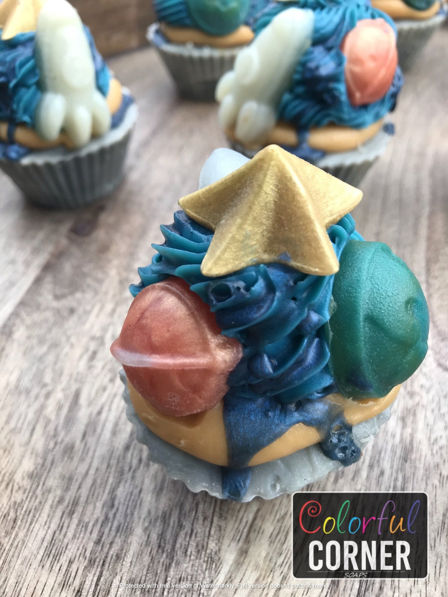 Again a new angle of upright zoomed in cupcakes. Shows cupcake side with Earth embed and Saturn embeds with star embed on top. Cupcake bottom is light and dark gray. Frosting is bright yellow ring on top of cupcake base then dark blue frosting in a swirl ring on top of that yellow ring. 