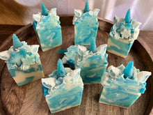 Load image into Gallery viewer, Snowflake the Magical Unicorn Bar On Display wooden starving platter. Snowflake the Magical Unicorn Bar front and center zoomed in on single bar of soap. Shows crystal like inside from salt and swirls of light blue teal and white. Frosted blue unicorn horn has a beautiful rough texture like a frosted glass. Ears are white with teal painted insides. Sides of unicorn frosted soap bar top are covered in snowflake imbeds.
