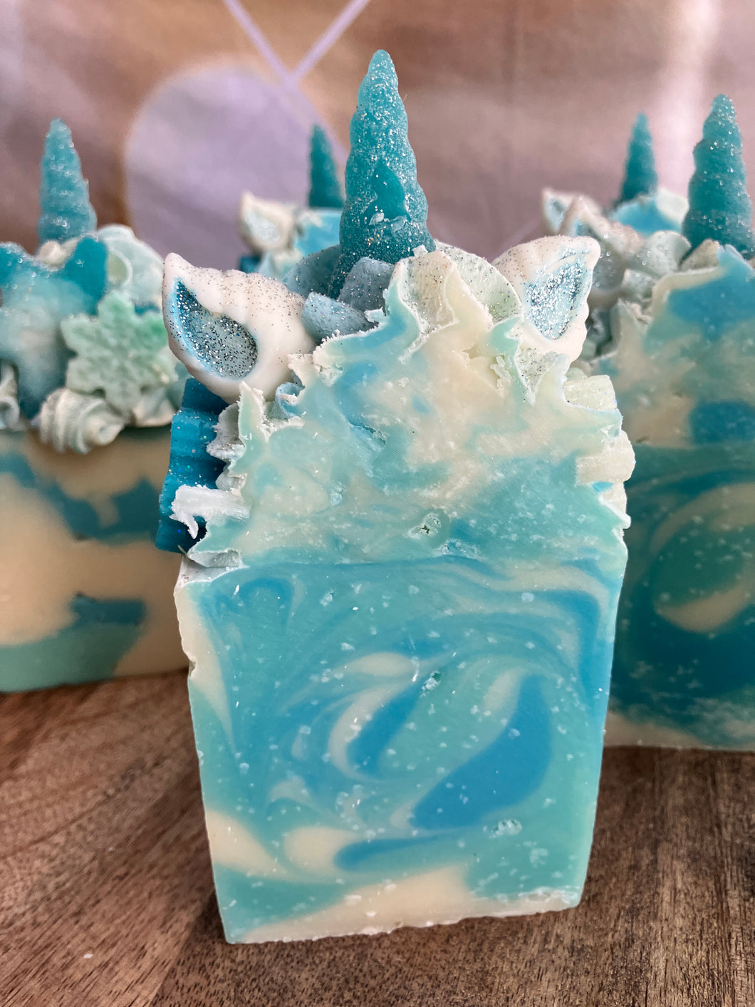 Snowflake the Magical Unicorn Bar front and center zoomed in on single bar of soap. Shows crystal like inside from salt and swirls of light blue teal and white. Frosted blue unicorn horn has a beautiful rough texture like a frosted glass. Ears are white with teal painted insides. Sides of unicorn frosted soap bar top are covered in snowflake imbeds.