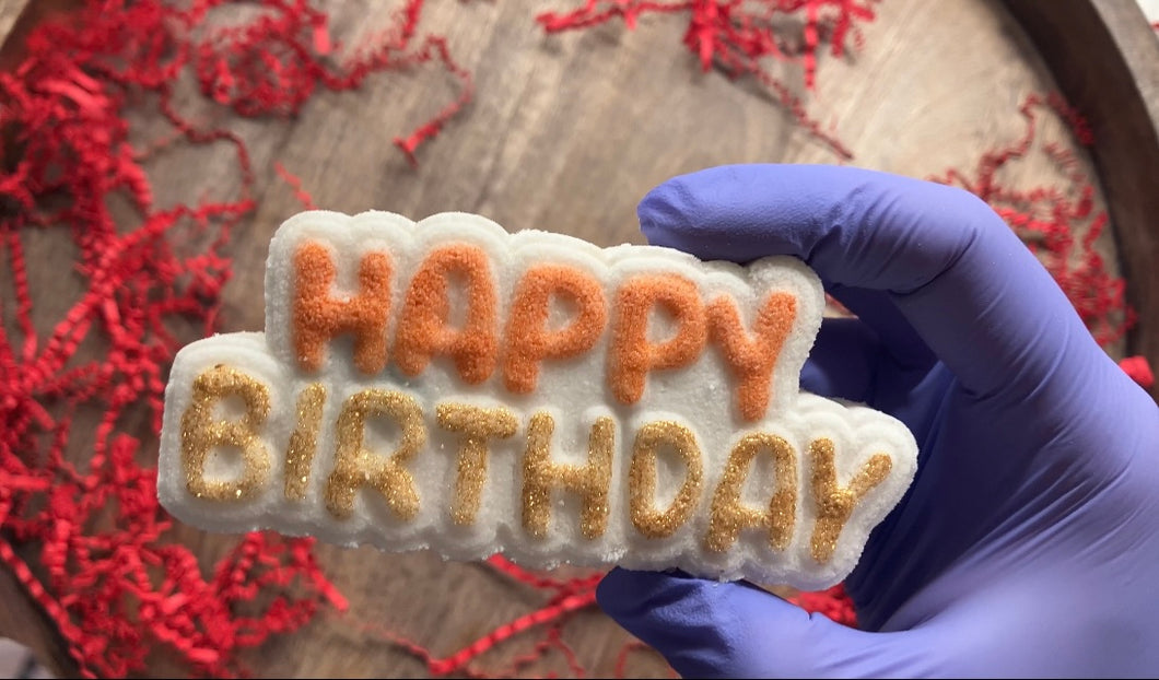 Close up of the 'Happy Birthday' bath bomb. Happy is orange and Birthday is gold.